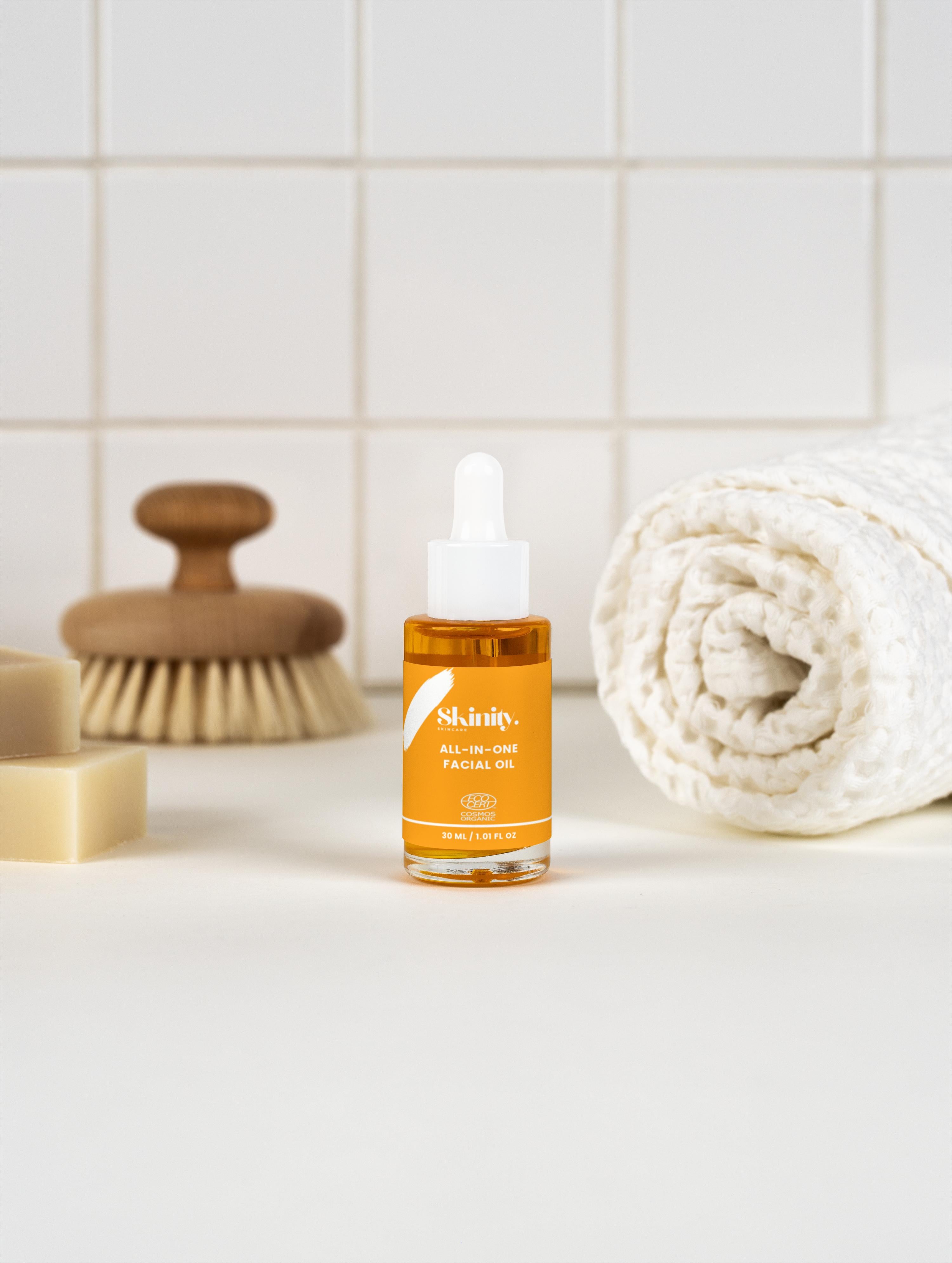 All-In-One Facial Oil for all skin in a stylish bottle beside rolled towel and cleaning brush.
