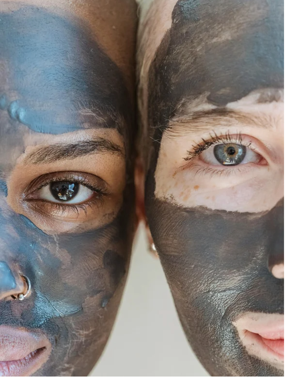 Close-up of two faces side-by-side wearing a dark grey clay mask, focusing on the eyes, mask texture, and application areas.