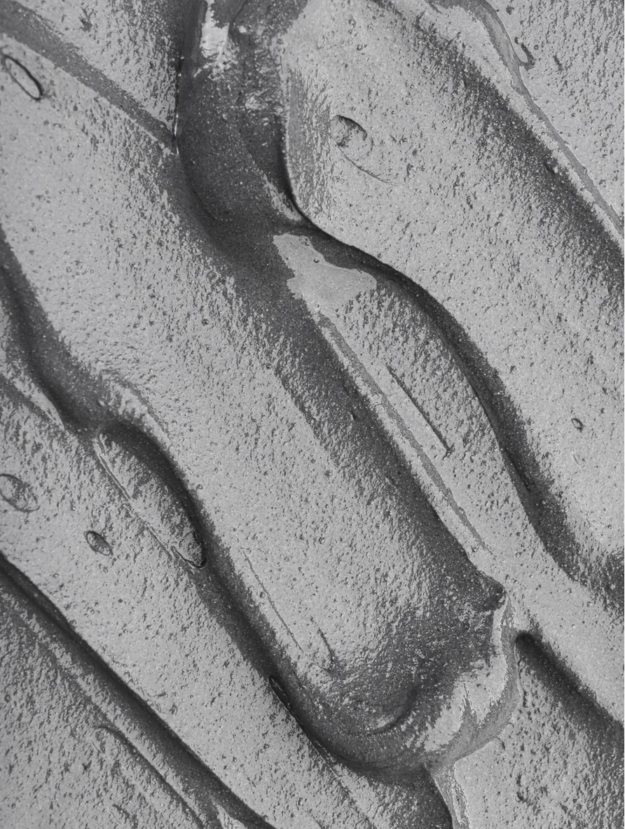 Macro close-up of dark grey clay mask texture with thick, creamy swirls and visible mineral particles on a flat surface.