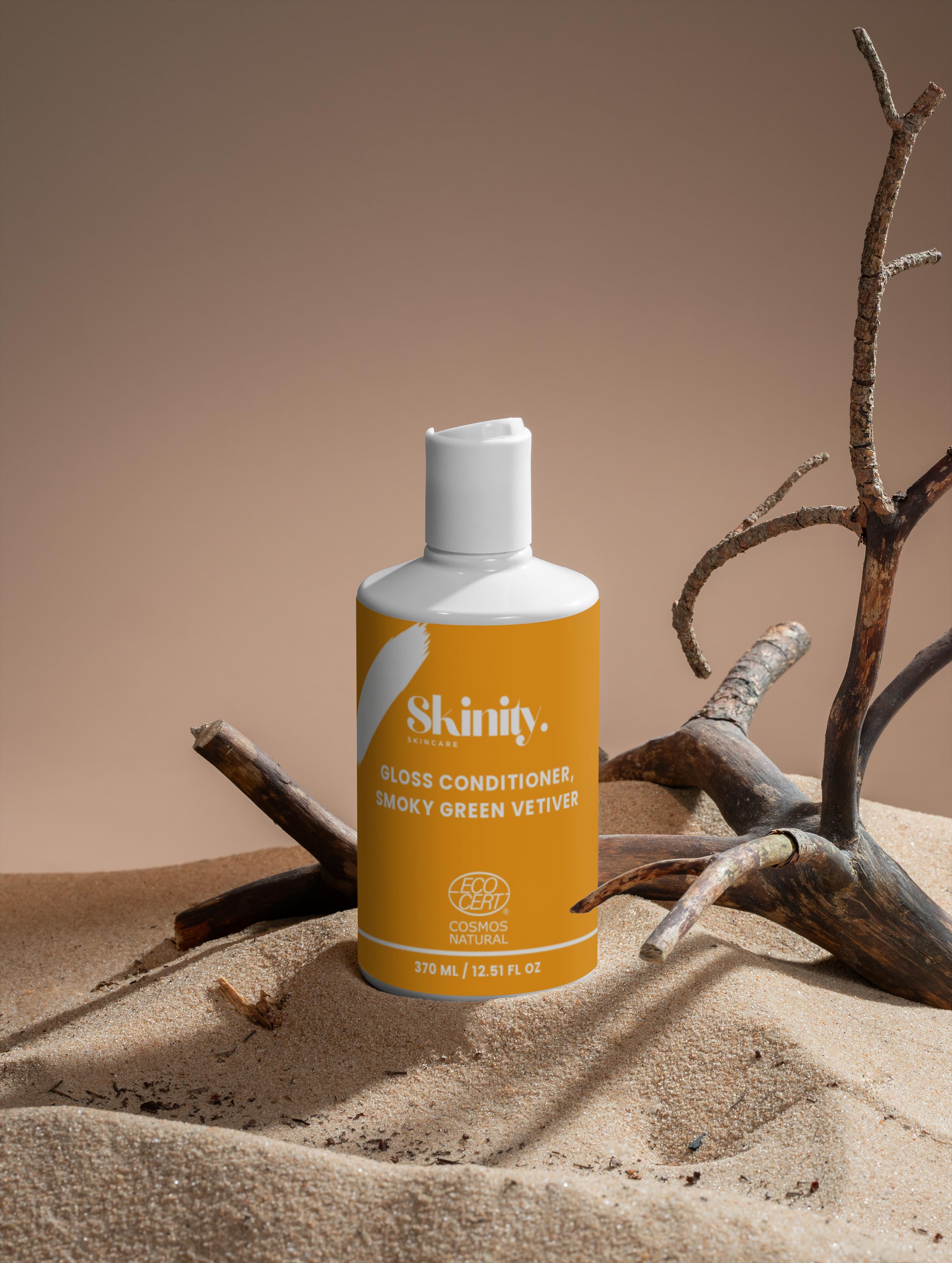 Gloss Conditioner Smoky Green Vetiver bottle displayed on sandy surface with twigs in the background.